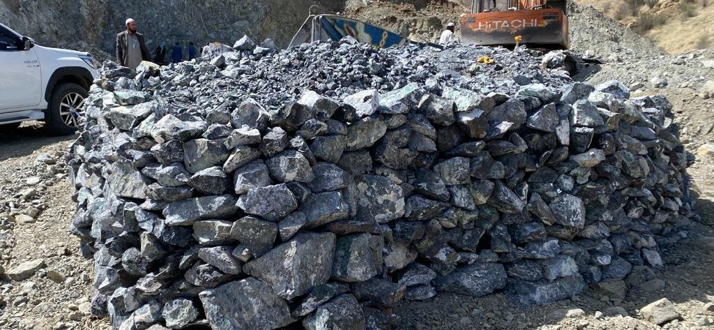 Pile of extracted metal ores at a Bare Syndicate mining site, with workers and machinery in the background.