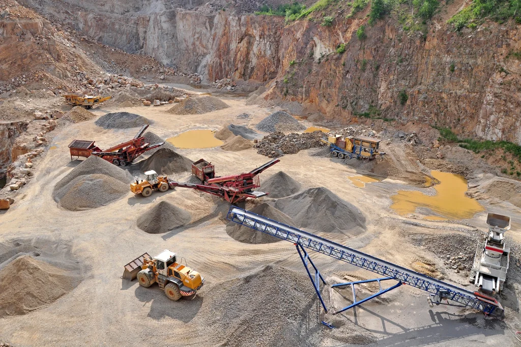 An aerial view of an active metal ore mining site with heavy machinery and conveyor belts processing extracted materials, illustrating the impact of economic shifts on mining operations.