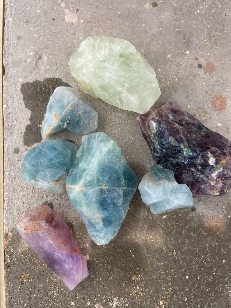 A collection of colorful fluorite mineral rocks in shades of green, blue, purple, and clear displayed on a concrete surface. These fluorite samples, used in various industrial applications, are sourced by Bare Syndicate.
