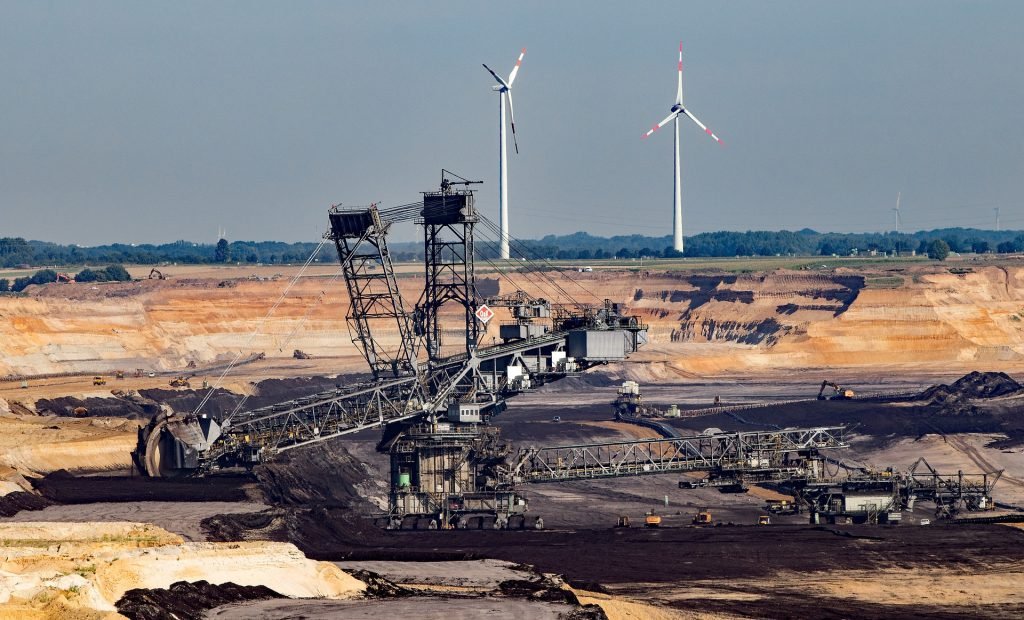 Mining operation with large excavators in an open-pit mine, with wind turbines in the background symbolizing the intersection of traditional and renewable energy.