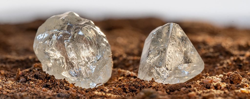 Two rough, uncut diamonds resting on a dirt surface, showcasing their natural, raw form.