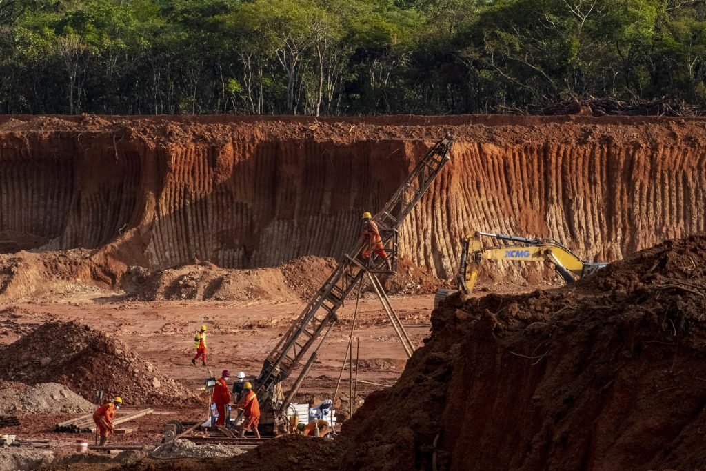 Workers conducting mining exploration at an open-pit mine, with equipment and excavators operating in a large reddish-brown mining site.