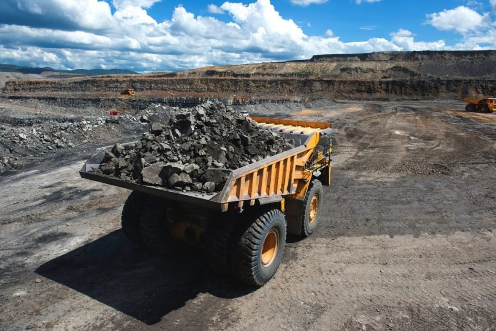 A large mining dump truck transporting rocks in an open-pit mining site under a clear blue sky with other machinery in the background.
