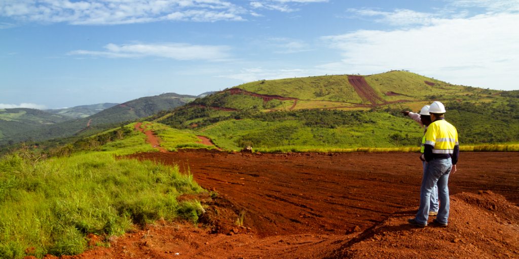 Two mining workers in safety gear surveying a rehabilitated mining site with restored vegetation and rolling green hills in the background under a bright blue sky.