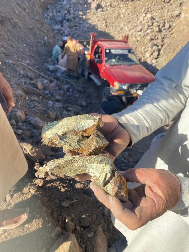 A miner holds freshly extracted high-grade copper ore at a mining site, highlighting the raw materials essential for the global copper supply. In the background, workers and a red transport truck facilitate the extraction and logistics. Bare Syndicate ensures the ethical sourcing and reliable distribution of premium-quality copper for industrial use.